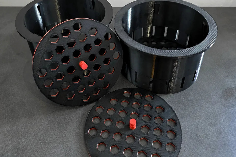 Black 3D printed filter cups with hexagon hole lids and red knobs on a grey surface