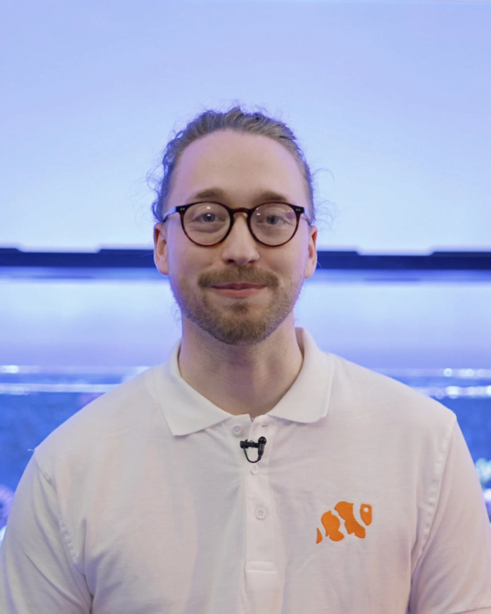 Harry from Marine Spec smiling in a white polo shirt with the clownfish logo, in front of a blue lit reef tank